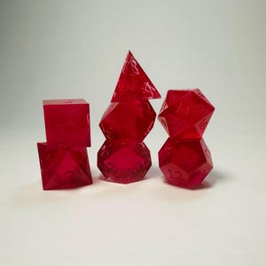 May include: A set of seven translucent, vibrant red polyhedral dice arranged in three stacks. The dice feature sharp edges and clearly engraved numbers. The collection includes various geometric shapes, including a cube, a pyramid, and icosahedrons.