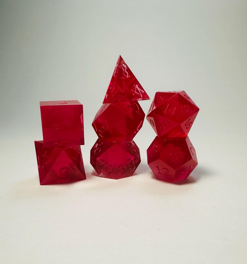 May include: A set of seven translucent, vibrant red polyhedral dice arranged in three stacks. The dice feature sharp edges and clearly engraved numbers. The collection includes various geometric shapes, including a cube, a pyramid, and icosahedrons.