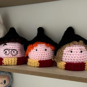 May include: Three crocheted plush figures wearing black pointed hats and scarves with alternating red and yellow stripes. The figures have light pink faces, black bead eyes, and yarn hair in dark brown, orange, and light brown colours.