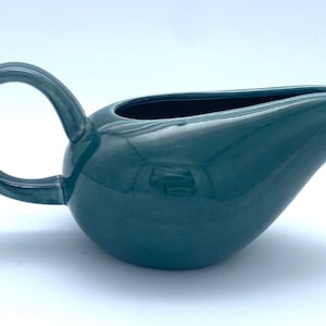 May include: A dark teal ceramic gravy boat with a smooth, glossy finish. The vessel features a rounded, bulbous body, a wide pouring spout, and a large, circular handle extending from the side, providing a functional grip for pouring liquids.
