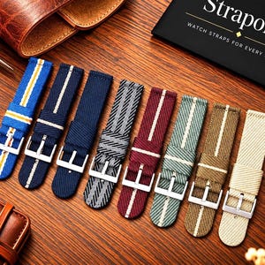 May include: A collection of eight fabric watch straps with silver-tone buckles, featuring various colours including blue, navy, grey, maroon, green, tan, and cream, some with striped patterns. A black card displays the text Straporia.