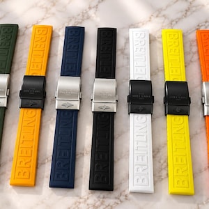 Könnte beinhalten: Sieben Kautschuk-Uhrenarmbänder in Olivgrün, Orange, Marineblau, Schwarz, Weiß, Gelb und leuchtendem Orange. Jedes Armband ist mit dem geprägten Schriftzug BREITLING und einer Metall-Faltschließe mit eingraviertem Breitling-Logo versehen.