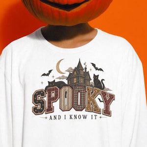 May include: A white long-sleeve shirt featuring a graphic of a haunted house, two black cats, bats, and a crescent moon. The word SPOOKY is printed in large, patterned block letters, with the text AND I KNOW IT centered underneath.
