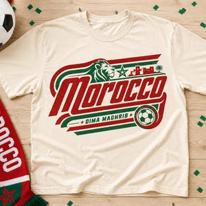 May include: A cream-colored short-sleeve t-shirt featuring a graphic with the text Morocco and Dima Maghrib, a lion head, a green star, a building silhouette, and a soccer ball in red and green colors, accompanied by a matching scarf.