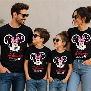May include: Four people wearing matching black t-shirts with a Minnie Mouse graphic and the text "Disney 2026". The Minnie Mouse design includes a pink bow, white gloves, and red accents. The family is wearing sunglasses and blue jeans.