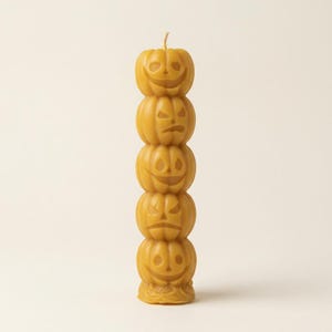 May include: A tall, cylindrical candle made of golden-yellow wax, featuring a vertical stack of five carved pumpkin faces. Each pumpkin displays a distinct facial expression, including smiling and frowning jack-o'-lantern designs.