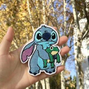 Può includere: Una toppa ricamata raffigurante un personaggio alieno azzurro con grandi orecchie e naso blu scuro, che tiene in mano una piccola rana verde con la pancia gialla. La toppa ha un bordo bianco e segue il contorno dei personaggi.