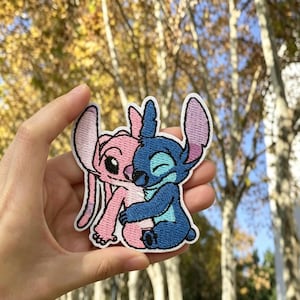 May include: An embroidered fabric patch featuring two cartoon characters in a close embrace. One character is light pink with large ears, while the other is dark blue with pointed ears and a closed-eye expression. The patch has a white border.