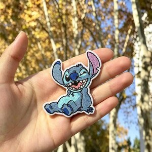 Puede incluir: Un parche bordado que presenta un personaje alienígena azul con orejas grandes, una amplia sonrisa que muestra los dientes y una barriga azul claro. El personaje está representado sentado con un borde blanco, mostrando un detallado trabajo de hilo.