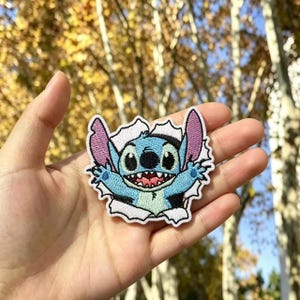 Könnte beinhalten: Ein gestickter Aufnäher, der den blauen Alien-Charakter Stitch mit großen Ohren, schwarzen Augen und einem breiten Grinsen zeigt, dargestellt als würde er durch einen weißen, zerrissenen Papierhintergrund mit gezackten Rändern brechen.