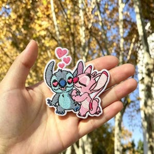 Può includere: Una toppa ricamata termoadesiva raffigurante un personaggio alieno azzurro con occhi a forma di cuore e un personaggio rosa con gli occhi chiusi, entrambi che si abbracciano. Tre piccoli cuori rosa fluttuano sopra, bordati in filo bianco.