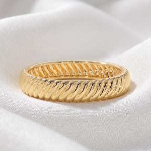 Puede incluir: Un anillo de oro que presenta una textura continua de cuerda o cable trenzado alrededor de toda la banda exterior. El metal tiene un acabado pulido y reflectante que capta la luz, creando una serie de crestas y surcos diagonales.