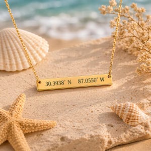 May include: A gold-colored necklace with a rectangular pendant engraved with coordinates. The necklace is displayed on a sandy surface with seashells and a starfish. The coordinates read 30.3938° N 87.0550° W.