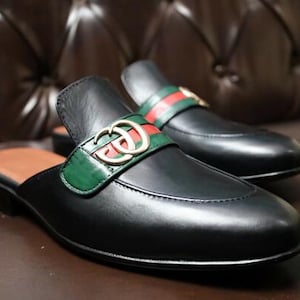 May include: Black leather slip-on shoes with a green and red accent strap and gold-coloured hardware. The shoes have a brown insole and a low heel. The shoes are displayed on a brown surface.