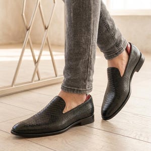 May include: Black leather slip-on shoes with a snakeskin texture. The shoes have a low heel and a pointed toe. The interior lining is red. The shoes are worn with grey jeans.