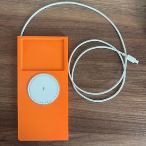 May include: Bright orange wireless charger with a white charging pad and a white cable. The charging pad has a lightning bolt symbol. The charger is rectangular with a recessed area at the top. The cable is coiled and has a USB-C connector.
