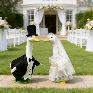 Könnte beinhalten: Zwei Gänsefiguren, die für eine Hochzeit gekleidet sind. Eine Gans trägt einen schwarzen Smoking mit Fliege und Zylinder. Die andere trägt ein weißes Brautkleid und einen Schleier. Sie stehen auf grünem Gras vor einem Hochzeitsort.