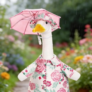 May include: A white goose plush toy wearing a pink floral raincoat, matching hat, and holding a small pink umbrella. The goose has a yellow beak and is set against a blurred garden background, creating a whimsical scene.