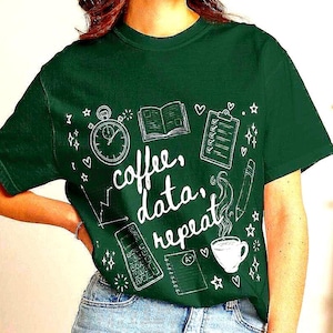 Può includere: Una maglietta verde scuro con la scritta bianca "coffee, data, repeat". La maglietta presenta illustrazioni bianche in stile doodle di un cronometro, un libro, un blocco note, una matita, una calcolatrice e una tazza di caffè. La modella indossa pantaloncini di jeans.