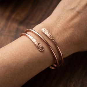 Op de afbeelding: Een koperen bypass-armband met een dubbel wikkelontwerp. Het metaal heeft een gepolijste afwerking met een duidelijke gehamerde textuur op de taps toelopende uiteinden, wat zorgt voor een reflecterend oppervlak op de gebogen metalen banden.