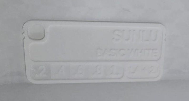 K&ouml;nnte beinhalten: Ein wei&szlig;es rechteckiges Objekt mit abgerundeten Ecken, mit dem gepr&auml;gten Text "SUNILU BASIC WHITE". Darunter befinden sich Zahlen und Symbole. Das Objekt hat ein kleines, erhabenes kreisf&ouml;rmiges Element.