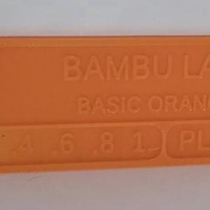 K&ouml;nnte beinhalten: Ein orangefarbenes rechteckiges Kunststoffobjekt mit abgerundeten Ecken. Auf der Oberfl&auml;che ist der Text "BAMBU LAB BASIC ORANGE" aufgedruckt, zusammen mit Zahlen und den Buchstaben "PLA". Oben links befindet sich ein kleines Loch.