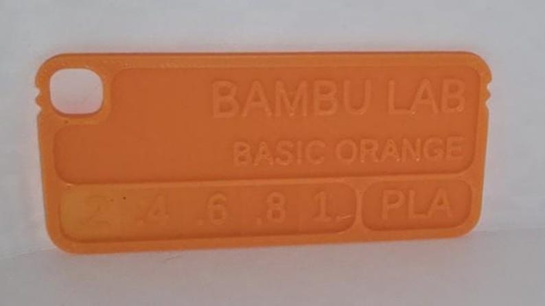 K&ouml;nnte beinhalten: Ein orangefarbenes rechteckiges Kunststoffobjekt mit abgerundeten Ecken. Auf der Oberfl&auml;che ist der Text "BAMBU LAB BASIC ORANGE" aufgedruckt, zusammen mit Zahlen und den Buchstaben "PLA". Oben links befindet sich ein kleines Loch.