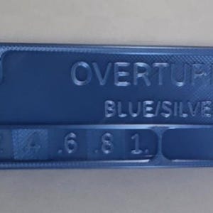 K&ouml;nnte beinhalten: Ein blaues rechteckiges Objekt mit abgerundeten Ecken, das den gepr&auml;gten Text "OVERTURE BLUE/SILVER" und numerische Markierungen aufweist. Das Objekt hat an einem Ende einen Ausschnitt und scheint ein Werkzeug oder eine Messlehre zu sein.