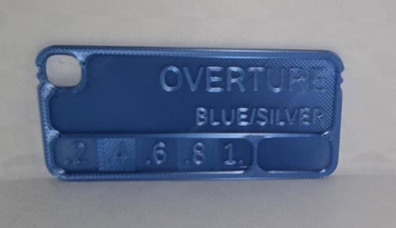 K&ouml;nnte beinhalten: Ein blaues rechteckiges Objekt mit abgerundeten Ecken, das den gepr&auml;gten Text "OVERTURE BLUE/SILVER" und numerische Markierungen aufweist. Das Objekt hat an einem Ende einen Ausschnitt und scheint ein Werkzeug oder eine Messlehre zu sein.