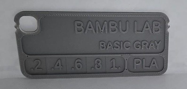 K&ouml;nnte beinhalten: Ein graues 3D-gedrucktes Farbmuster von Bambu Lab, beschriftet mit "Basic Gray". Das Muster enth&auml;lt Zahlen von .2 bis 1.0 und die Buchstaben "PLA". Der Artikel hat einen Ausschnitt auf der linken Seite.