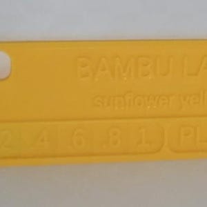 K&ouml;nnte beinhalten: Ein hellgelbes Kunststoff-Farbmuster mit abgerundeten Ecken und einem Loch. Der Text "BAMBU LAB sunflower yellow" ist auf der Oberfl&auml;che gepr&auml;gt, zusammen mit den Zahlen "2 4 6 8 1" und "PLA".