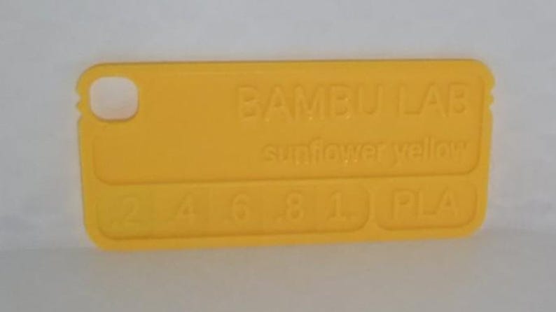 K&ouml;nnte beinhalten: Ein hellgelbes Kunststoff-Farbmuster mit abgerundeten Ecken und einem Loch. Der Text "BAMBU LAB sunflower yellow" ist auf der Oberfl&auml;che gepr&auml;gt, zusammen mit den Zahlen "2 4 6 8 1" und "PLA".