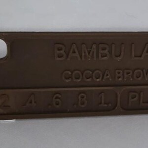 K&ouml;nnte beinhalten: Ein rechteckiges Farbmuster aus kakaobraunem PLA-Kunststoff von Bambu Lab. Das Muster hat abgerundete Kanten und ein kleines Loch an einem Ende. Der Text "BAMBU LAB COCOA BROWN" ist zusammen mit Zahlen und den Buchstaben "PLA" gepr&auml;gt.