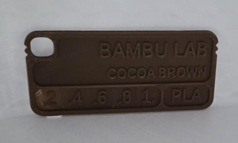 K&ouml;nnte beinhalten: Ein rechteckiges Farbmuster aus kakaobraunem PLA-Kunststoff von Bambu Lab. Das Muster hat abgerundete Kanten und ein kleines Loch an einem Ende. Der Text "BAMBU LAB COCOA BROWN" ist zusammen mit Zahlen und den Buchstaben "PLA" gepr&auml;gt.