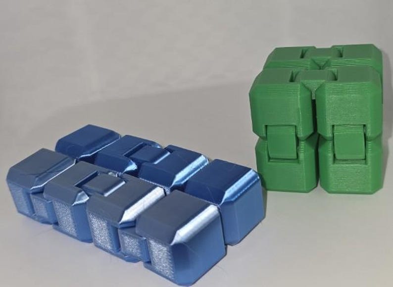 K&ouml;nnte beinhalten: Gezeigt werden zwei Zappelspielzeuge. Eines ist ein blauer Infinity-W&uuml;rfel, das andere ein gr&uuml;ner Infinity-W&uuml;rfel. Der blaue W&uuml;rfel besteht aus mehreren verbundenen Bl&ouml;cken. Der gr&uuml;ne W&uuml;rfel besteht aus vier verbundenen Bl&ouml;cken. Beide befinden sich auf einer wei&szlig;en Oberfl&auml;che.
