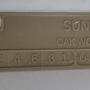 K&ouml;nnte beinhalten: Ein beigefarbenes rechteckiges Objekt mit abgerundeten Ecken. Der Text "SUNLU OAK WOOD" ist auf der Oberfl&auml;che gepr&auml;gt. Darunter sind Zahlen von .2 bis 1 gedruckt, gefolgt von "PLA + 2.0". Das Objekt hat ein kleines Loch in der oberen linken Ecke.