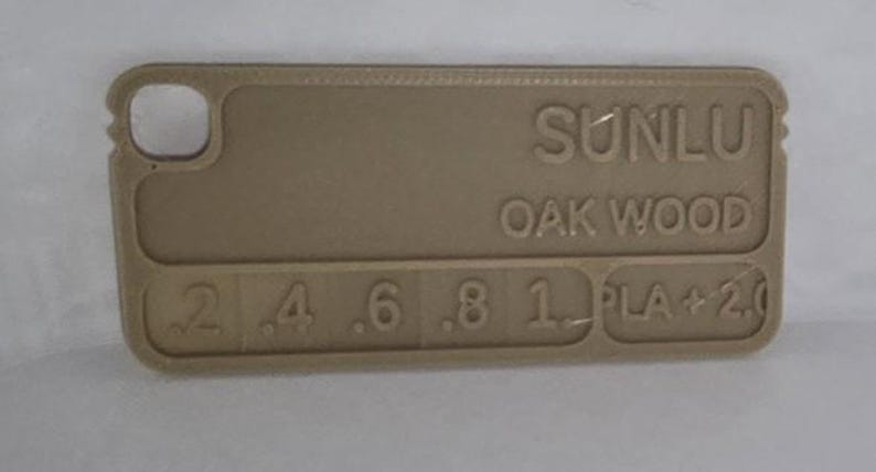 K&ouml;nnte beinhalten: Ein beigefarbenes rechteckiges Objekt mit abgerundeten Ecken. Der Text "SUNLU OAK WOOD" ist auf der Oberfl&auml;che gepr&auml;gt. Darunter sind Zahlen von .2 bis 1 gedruckt, gefolgt von "PLA + 2.0". Das Objekt hat ein kleines Loch in der oberen linken Ecke.