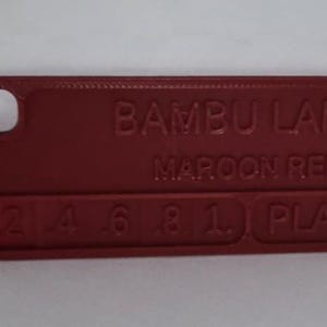 K&ouml;nnte beinhalten: Maroonrotes rechteckiges Kunststoff-Farbmuster mit abgerundeten Ecken und einem Loch. Der Text "BAMBU LAB MAROON RED" ist auf der Oberfl&auml;che gepr&auml;gt, zusammen mit Zahlen und "PLA".