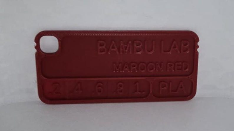 K&ouml;nnte beinhalten: Maroonrotes rechteckiges Kunststoff-Farbmuster mit abgerundeten Ecken und einem Loch. Der Text "BAMBU LAB MAROON RED" ist auf der Oberfl&auml;che gepr&auml;gt, zusammen mit Zahlen und "PLA".