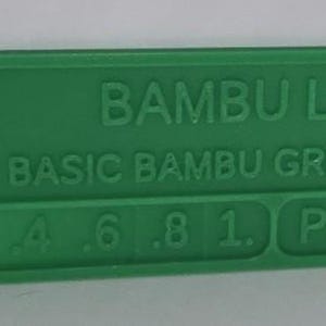 K&ouml;nnte beinhalten: Ein gr&uuml;nes Kunststoff-Farbmuster von Bambu Lab, beschriftet mit "Basic Bambu Green". Das Muster enth&auml;lt den Text "PLA" und Zahlen von .2 bis 1. Es hat ein kleines Loch an einem Ende.