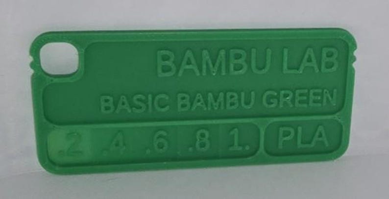 K&ouml;nnte beinhalten: Ein gr&uuml;nes Kunststoff-Farbmuster von Bambu Lab, beschriftet mit "Basic Bambu Green". Das Muster enth&auml;lt den Text "PLA" und Zahlen von .2 bis 1. Es hat ein kleines Loch an einem Ende.