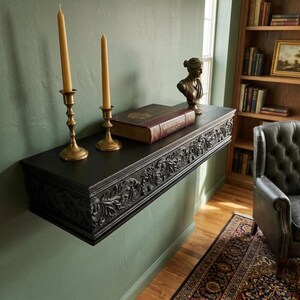 May include: A dark, carved shelf fixed to a green wall, with two brass candlesticks holding lit candles, a book, and a bust. The shelf has detailed carvings along the front. A bookcase and armchair are in the background.