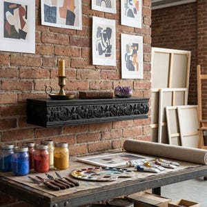 May include: An artist's studio featuring a brick wall with framed abstract art. A black, carved shelf holds a candle and a crystal. A table displays paint jars, a palette, paint tubes, and a canvas roll. An easel and canvases are in the background.