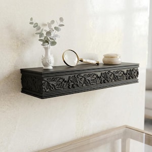 May include: A black, rectangular wall shelf with detailed floral carvings. A small white vase with foliage, a magnifying glass, and a small decorative jar are on the shelf. The shelf is mounted on a cream-coloured wall.