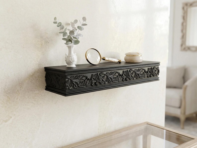 May include: A black, rectangular wall shelf with detailed floral carvings. A small white vase with foliage, a magnifying glass, and a small decorative jar are on the shelf. The shelf is mounted on a cream-coloured wall.