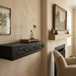 May include: A dark, carved wooden shelf with floral detailing is fixed to a beige wall. A framed botanical print and a small glass vase with dried flowers are placed above the shelf. A framed painting hangs on the wall.