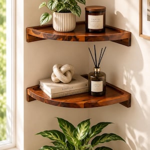 May include: Two dark brown wooden corner shelves mounted on a wall. The top shelf holds a green plant in a white ribbed pot and a brown candle labeled Calming Atmosphere. The bottom shelf holds a beige knot sculpture, two books, and a reed diffuser.
