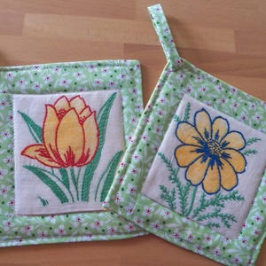 Puede incluir: Dos agarraderas cuadradas con bordados florales. Una presenta un tulipán amarillo y rojo, la otra una margarita amarilla y azul. Ambas están enmarcadas con una tela floral verde claro y tienen un lazo para colgar.