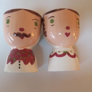 May include: Two ceramic egg cups, one depicting a man with a mustache and the other a woman with a heart. The man's egg cup has a red and green patterned bow tie, while the woman's has a red and green scalloped neckline.