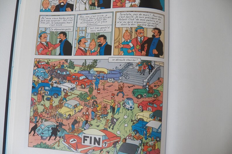 May include: A comic book page featuring Tintin characters in dialogue panels above a large, detailed illustration of a car rally scene. The scene depicts numerous vintage cars in various colors, people, and a finish line sign labeled FIN.